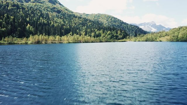 Aerial view; drone flying low over emerald water surfase of lake surrounded by mountain ranges and valleys; reflecting of lakesides on water; Agepsta mountain range with glaciers on background