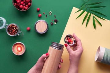 Zero waste tea to go, making herbal infusion in eco friendly insulated bamboo steel flask with herbal mixture and fresh cranberry. Trendy creative flat lay, top view on two tone green yellow paper.