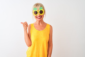 Middle age woman on vacation wearing pineapple sunglasses over isolated white background smiling with happy face looking and pointing to the side with thumb up.