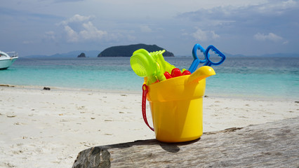 Colorful kid sand scoop toy in yellow plastic bucket on the timber log with beautiful beach background