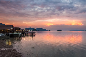 Fototapeta na wymiar Beautiful sunset in Thailand on Koh Lanta. Along the shore are houses with evening illumination. On the horizon are mountain hills. The looming cloudy sky is reflected in the calm sea