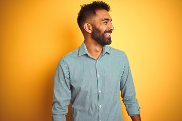 Young indian man wearing green striped shirt standing over isolated yellow background looking away...