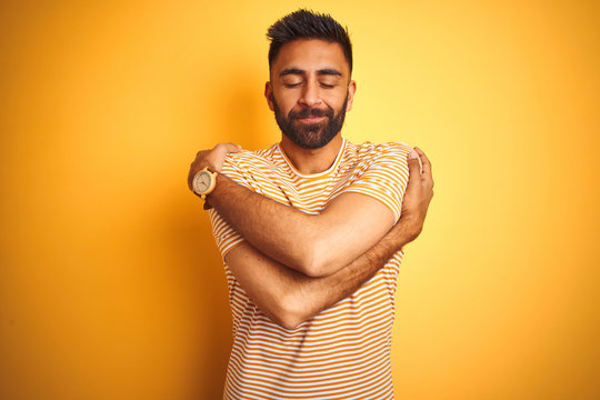 Young indian man wearing t-shirt standing over isolated yellow background Hugging oneself happy and positive, smiling confident. Self love and self care