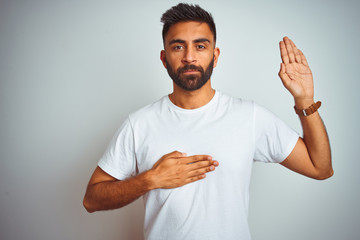 Young indian man wearing t-shirt standing over isolated white background Swearing with hand on...