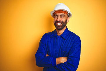 Handsome indian worker man wearing uniform and helmet over isolated yellow background happy face...