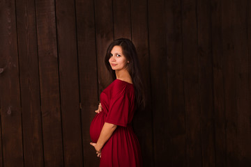 Fototapeta premium Pregnant woman in red dress holds hands on belly on a dark brown background. Pregnancy, maternity, expectation concept. Beautiful tender mood photo of pregnancy
