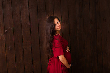 Fototapeta premium Pregnant woman in red dress holds hands on belly on a dark brown background. Pregnancy, maternity, expectation concept. Beautiful tender mood photo of pregnancy