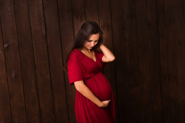 Fototapeta premium Pregnant woman in red dress holds hands on belly on a dark brown background. Pregnancy, maternity, expectation concept. Beautiful tender mood photo of pregnancy