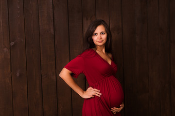 Pregnant woman in red dress holds hands on belly on a dark brown background. Pregnancy, maternity, expectation concept. Beautiful tender mood photo of pregnancy