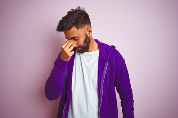 Young indian man wearing purple sweatshirt standing over isolated pink background tired rubbing nose and eyes feeling fatigue and headache. Stress and frustration concept.