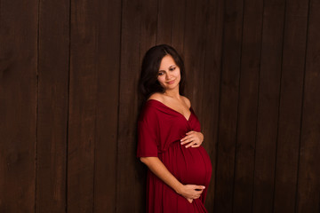 Fototapeta premium Christmas pregnant woman in red dress holds hands on belly on a dark brown background. Pregnancy, maternity, expectation concept. Beautiful tender mood photo of pregnancy