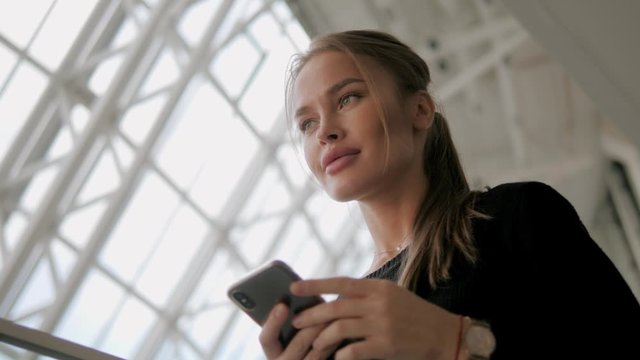 Attractive blonde young woman with big full sexy lips using smart phone. Pretty lady communicating by telephone, reads a message and smiles inside modern business center. Slow motion, close up.