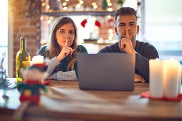 Young beautiful couple sitting using laptop around christmas decoration at home asking to be quiet with finger on lips. Silence and secret concept.