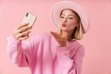 Portrait of woman in hat blowing air kiss at camera while taking selfie