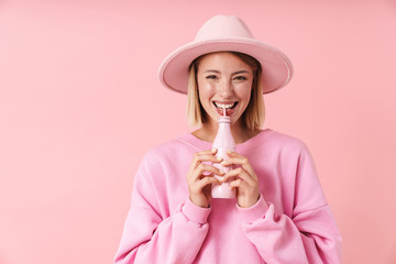 Portrait of optimistic woman in hat smiling while drinking from bottle