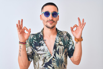 Young handsome man wearing Hawaiian sumer shirt and blue sunglasses over isolated background relax and smiling with eyes closed doing meditation gesture with fingers. Yoga concept.