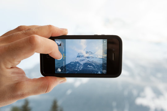 GRINDELWALD, SWITZERLAND - FEBRUARY 4, 2014: Man taking a photo of the Eiger mountain using the camera app on an Apple iPhone 4s. Close-up shot of the hand holding the phone with the mountain behind.