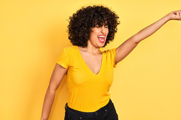 Young arab woman with curly hair wearing t-shirt standing over isolated yellow background Dancing happy and cheerful, smiling moving casual and confident listening to music