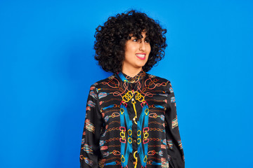 Young arab woman with curly hair wearing colorful shirt over isolated blue background looking away to side with smile on face, natural expression. Laughing confident.