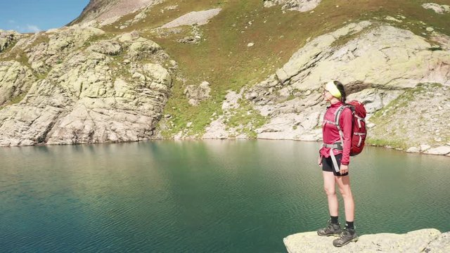 Young brunette hiker girl standing on stony cliff edge, looking at mountain landscape, beautiful highland lake, crystal clear turquoise water, sky reflection; travaler with backpack in climbing boots