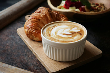 A cup of coffee with latte art on top, butter  croissant and granola with fresh tropical fruits and yogurt on table in caffee. Concept of healthy breakfast