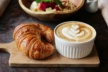 A cup of coffee with latte art on top, butter  croissant and granola with fresh tropical fruits and yogurt on table in caffee. Concept of healthy breakfast