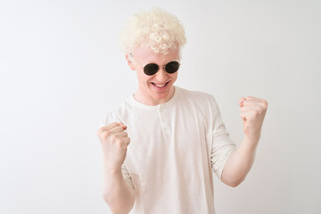 Young albino blond man wearing t-shirt and sunglasses over isolated white background very happy and excited doing winner gesture with arms raised, smiling and screaming for success. Celebration 