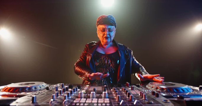 A cool caucasian dj grandma is composing a mix at turntables controller. Authentic mature woman working as disc jockey in a nightclub 4k footage