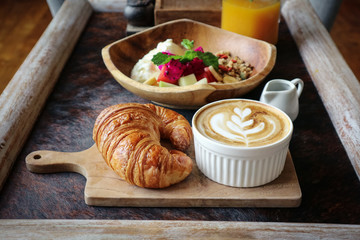A cup of coffee with latte art on top, butter  croissant and granola with fresh tropical fruits and yogurt on table in caffee. Concept of healthy breakfast