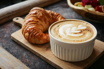 A cup of coffee with latte art on top, butter  croissant and granola with fresh tropical fruits and yogurt on table in caffee. Concept of healthy breakfast