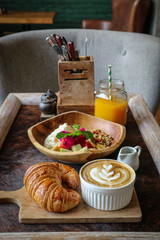A cup of coffee with latte art on top, butter  croissant and granola with fresh tropical fruits and yogurt on table in caffee. Concept of healthy breakfast