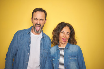 Beautiful middle age couple together wearing denim shirt over isolated yellow background sticking tongue out happy with funny expression. Emotion concept.