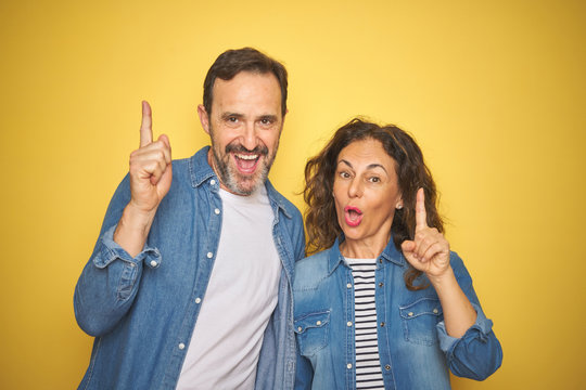 Beautiful middle age couple together wearing denim shirt over isolated yellow background pointing finger up with successful idea. Exited and happy. Number one.
