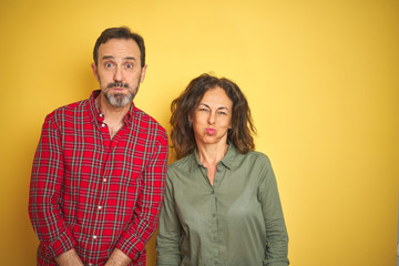 Beautiful middle age couple over isolated yellow background puffing cheeks with funny face. Mouth inflated with air, crazy expression.