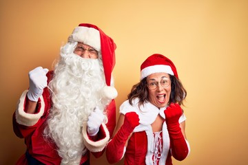 Middle age couple wearing Santa costume and glasses over isolated yellow background very happy and excited doing winner gesture with arms raised, smiling and screaming for success. Celebration concept