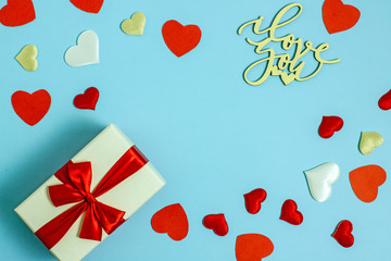 Valentines day background copy space. On a blue background a gift box and a red bow, red paper hearts, white fabric and red satin hearts and the inscription made of wooden letters, I love you