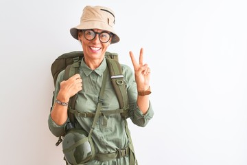 Fototapeta premium Middle age hiker woman wearing backpack canteen hat glasses over isolated white background smiling with happy face winking at the camera doing victory sign. Number two.