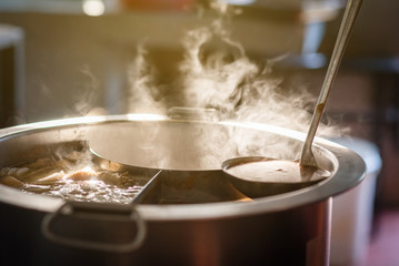 Cooking person in the restaurant is cooking while using the dipper in a large soup pot. And the water is boiling, there is smoke and hot spit.