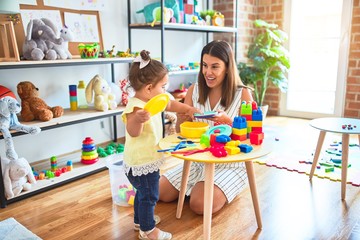 Young beautiful teacher and toddler playing with dishes, cutlery and cups toy on the table at kindergarten