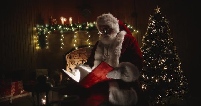 Authentic santa clause opening his magical shining book with red cover, sitting on rocker near fireplace and christmas tree - holiday mood, christmas spirit concept 4k footage
