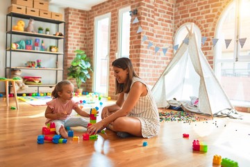 Beautiful teacher and blond toddler girl building tower using plastic blocks at kindergarten