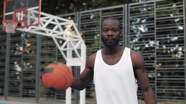 Portrait Muscly Handsome Afro - American Guy in White Singlet Dribbling a Ball Standing at Street Basketbal Sports Field and Looking to Camera. Healthy Lifestyle and Sport Concept.