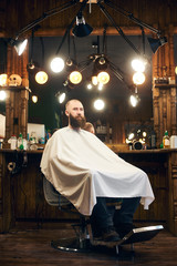 Adult client with beard after service sitting in hairdresser chair and waiting for master in barber shop with modern vintage wooden interior. Concept of professional male care. Vertical general view.