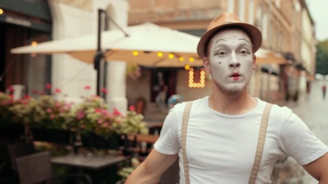 Young handsome mime is performing on street near cafe, denies, says no certainly. Performing by entertainer in hat, suspenders, makeup. Artist is moving arms, looks scared.