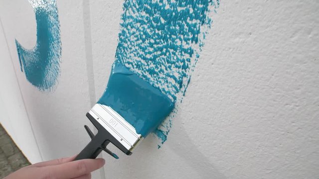 Close-up slow motion of a hand painting a blue letter on a white wall with a paint brush.  hand lubricating paint on a wall. blue being painted on the wall. Redecorating a white wall. Girls hand. A