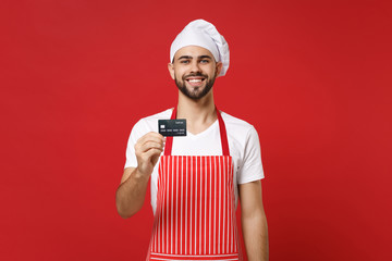 Smiling young bearded male chef cook or baker man in striped apron white t-shirt toque chefs hat isolated on red wall background. Cooking food concept. Mock up copy space. Holding credit bank card.