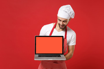 Funny young male chef cook or baker man in striped apron t-shirt toque chefs hat isolated on red background. Cooking food concept. Mock up copy space. Hold laptop pc computer with blank empty screen.