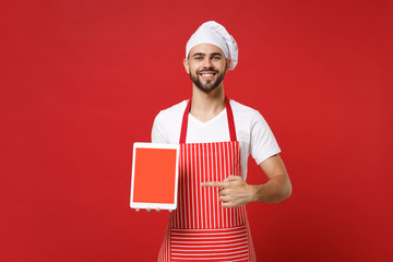 Smiling chef cook or baker man in striped apron toque chefs hat isolated on red background. Cooking food concept. Mock up copy space. Pointing index finger on tablet computer with blank empty screen. - Powered by Adobe