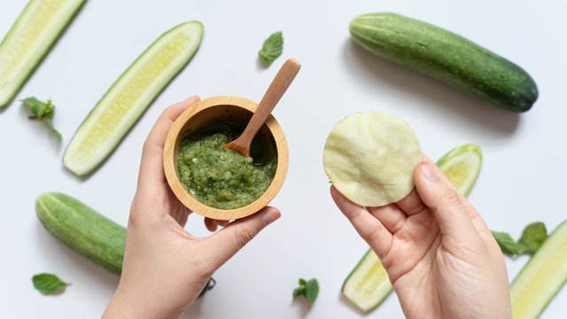 Cucumber mask or makeup remover pads made w/ natural ingredients for healthy skincare. Hands holding DIY eyes/facial mask and a bowl of cucumber mixed w/ mint on white table. Homemade beauty product.