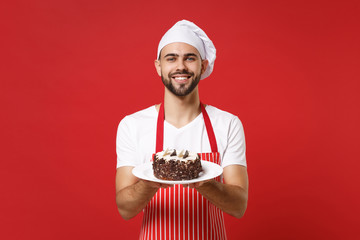 Smiling young bearded male chef cook or baker man in striped apron white t-shirt toque chefs hat posing isolated on red background. Cooking food concept. Mock up copy space. Holding plate with cake.